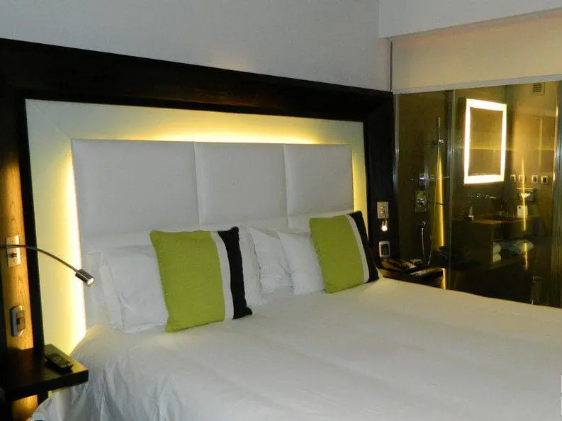 Uma das Suites Novotel, cama com iluminação e ao fundo box para banho de vidro