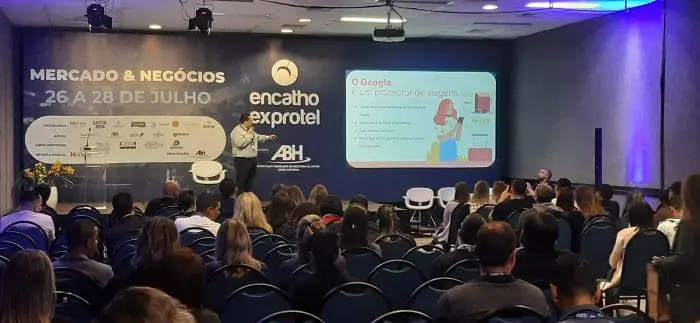 Diego Corrêa, CMO da HSystem, palestra sobre a geração de demanda hoteleira e até mesmo aérea, como é o caso do Google. Diego aponta para um projetor e fala sobre o Google na demanda aérea, no Encatho & Exprotel 2022, em Florianópolis-SC.