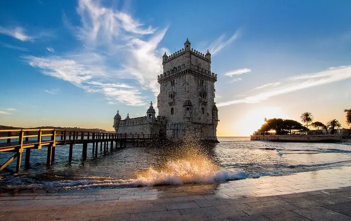 Vista do Jardim da Torre de Belém, em Lisboa, Portugal. Embaixo da Torre, há o mar, e uma onda quebrou próxima à calçada. Ao fundo, o céu azul com algumas nuvens e o sol de entardecer.