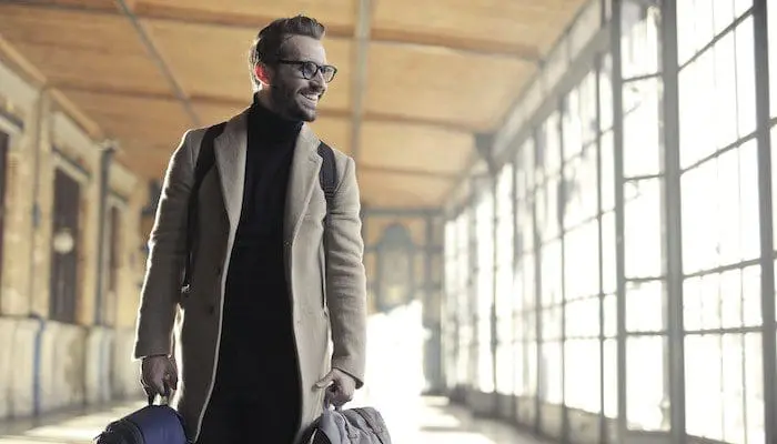 Homem loiro de óculos, usando sobretudo e segurando duas malas nas mãos e uma mochila nas costas, está sorrindo e fará uma de suas viagens a trabalho.