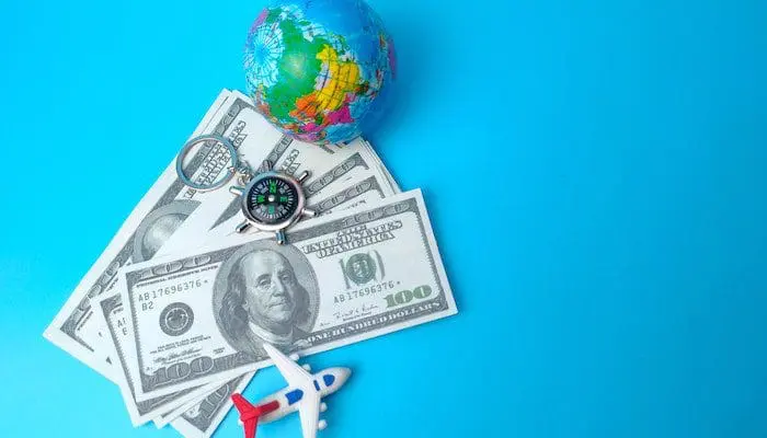 Imagem com fundo azul, com notas de dólar, bússola, globo pequeno e avião para falar sobre pedidos de empréstimo para viagens.