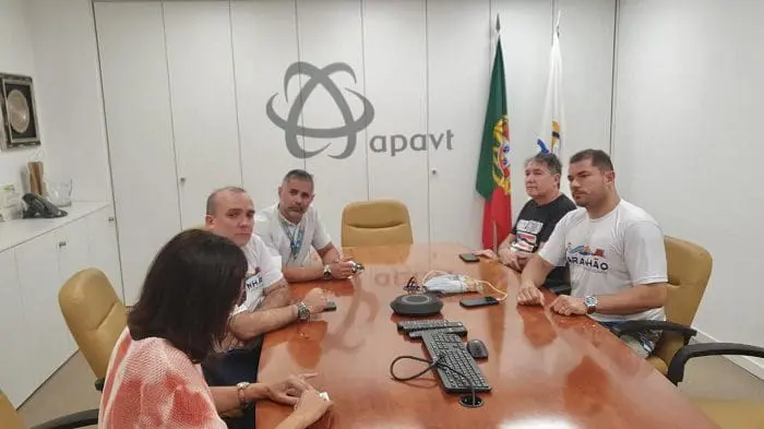 Representantes da SETUR São Luís se reúnem com representantes da da Associação Portuguesa das Agências de Viagens e Turismo (APAVT) no escritório da associação. Todos estão sentados à mesa, conversando. Ao fundo, está o logo da APAVT na parede e do lado direito, a bandeira de Portugal.