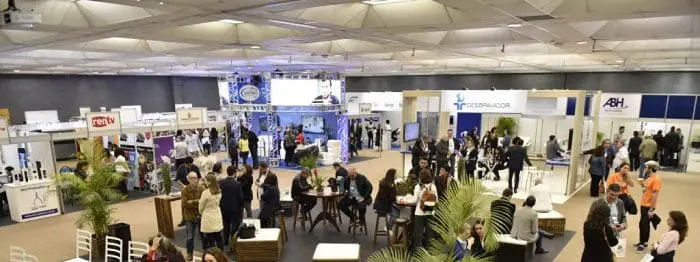 Evento Exprotel, com estandes de hotéis e pessoas circulando. Algumas pessoas estão sentadas, conversando.