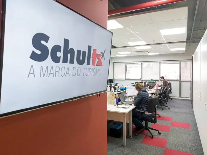 Entrada do escritório da Schultz, com uma TV do lado esquerdo que exibe o nome da empresa e o slogan, e ao fundo do escritório, do lado direito, os colaboradores da empresa estão sentados, trabalhando.