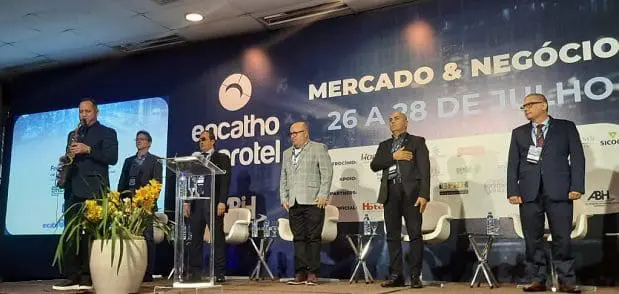 O Hino Nacional é entoado com saxofone por um saxofonista, à esquerda da foto; e autoridades participam da solenidade. Um dos presentes está com a mão direita no peito, em sinal de respeito. Atrás, o painel da Encatho & Exprotel, que é realizada em Florianópolis-SC.