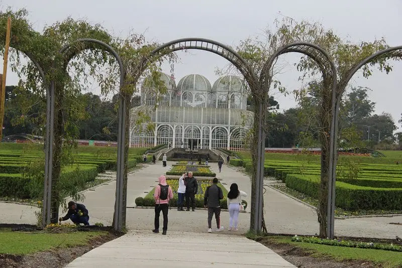 Foto de um portal que dá acesso ao jardim e à estufa do Jardim Botânico, em Curitiba, no Paraná. Embaixo do portal, estão algumas pessoas, que passam por ele para acessar o jardim.