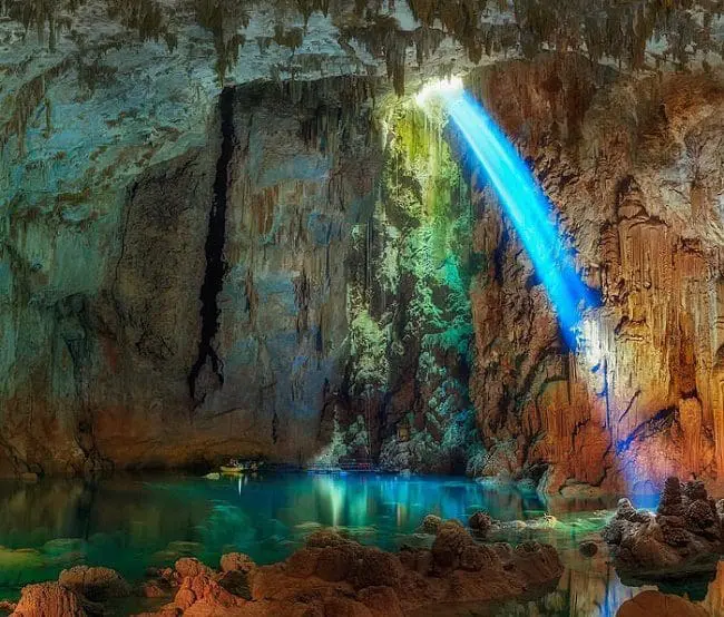 Vista de uma caverna de Bonito, no Mato Grosso do Sul. A caverna possui formações rochosas em tom marrom alaranjado, água em tons azul turquesa e verde, uma abertura superior, pela qual passa a luz do sol e reflete a luz azul turquesa na pedra.