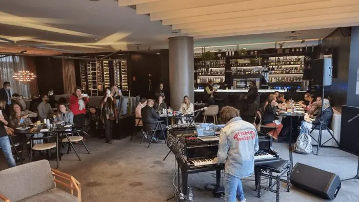 evento_rock_n_taste_it_pullman_ibirapuera Evento Rock´n Taste IT, no Pullman Ibirapuera. Há algumas pessoas comendo na mesa, um músico tocando piano, e outas pessoas, à esquerda, assistindo.