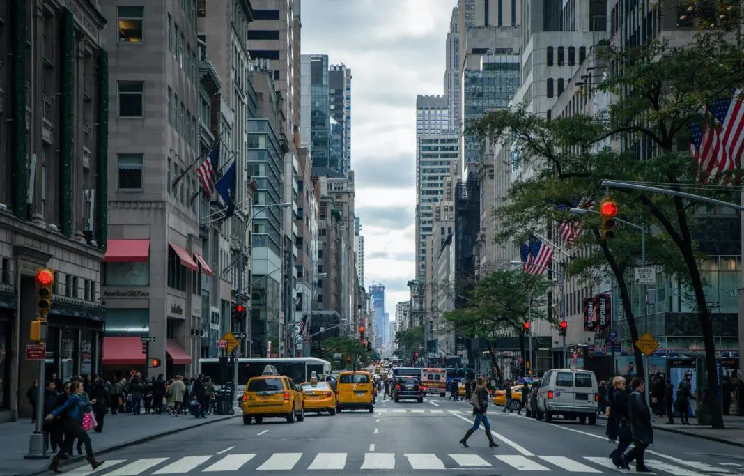 pexels-nout-gons-378570 Uma das ruas da cidade de Nova York, nos Estados Unidos. Há táxis amarelos no trânsito, prédios com bandeiras dos Estados Unidos hasteadas, e uma pessoa atravessando a rua.