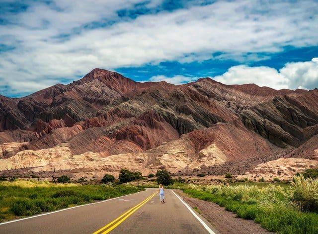 Estrada com uma pessoa de costas, caminhando, e ao fundo, montanhas altas de cor marrom avermelhado debaixo do céu azul com algumas nuvens, em em Salta, na Argentina.