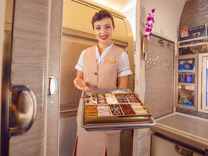 Comissária de bordo oferece, sorrindo, diversos aperitivos em uma bandeja a bordo de uma aeronave da Emirates.