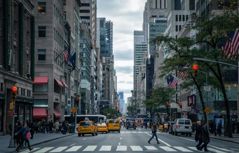 Uma das ruas da cidade de Nova York, nos Estados Unidos. Há táxis amarelos no trânsito, prédios com bandeiras dos Estados Unidos hasteadas, e uma pessoa atravessando a rua.