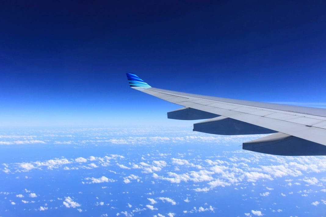 Vista da janela de uma asa de um avião, em pleno voo, sobrevoando as nuvens. O tom predominante do céu e das nuvens é azul claro.
