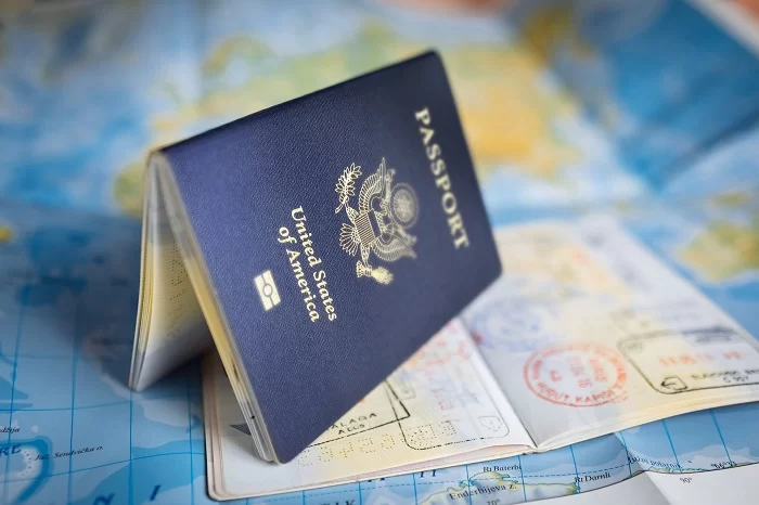 Foto de um passaporte americano aberto na vertical, para baixo. Ele está em cima de outro passaporte aberto, com vários carimbos, e os dois estão sobre um mapa mundi azul. Espera para renovação de visto americano de negócios e turismo diminui.