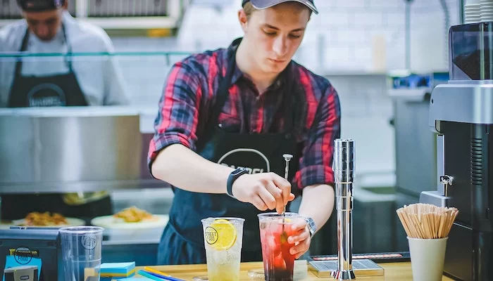 Garçom de boné usando camisa xadrez vermelha e preta e avental, mistura um drink vermelho em cima de um balcão de madeira clara. Do lado esquerdo, há outra bebida de cor branca, com uma laranja dentro. Empregos no Brasil: 1 em cada 5 criados em novembro foram do Turismo.