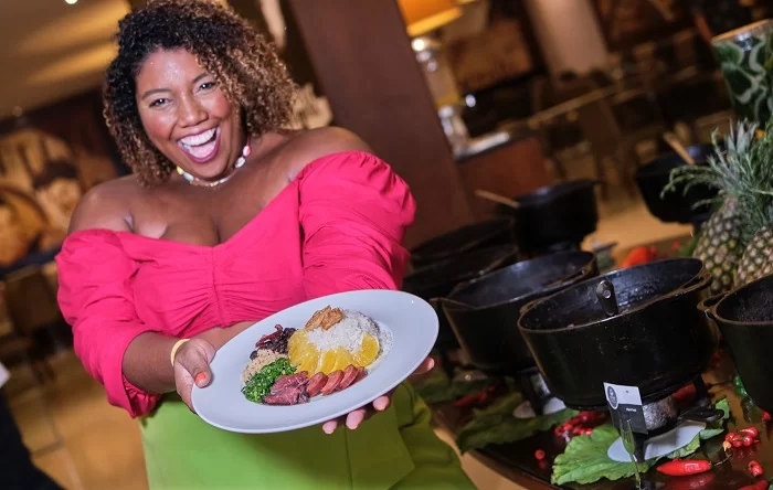 Mulher negra, sorridente e de blusa rosa, segura um prato com a feijoada pré-Carnavalesca do Vila Galé Rio de Janeiro. O evento acontece no dia 28 de janeiro.