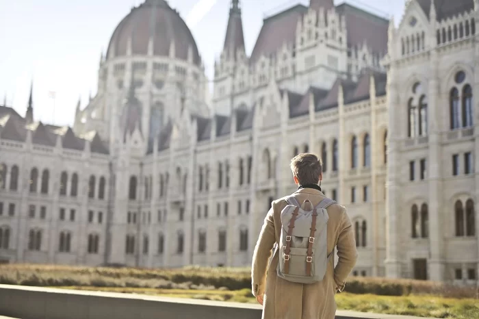 Homem de costas, com roupa social e mochila, vendo um castelo na Alemanha, de dia. Turismo na Alemanha: veja quanto se gasta com saúde em casos de emergência e a importância do seguro viagem