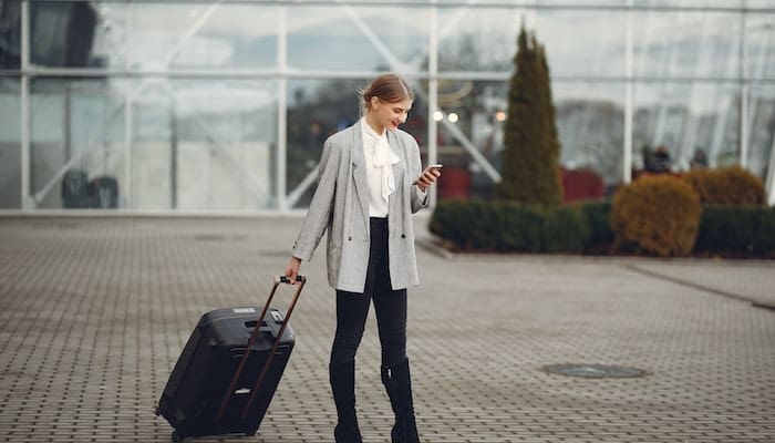 Viagens corporativas no 1º semestre: mulher vestida de social, leva mala de rodinhas e olha o celular