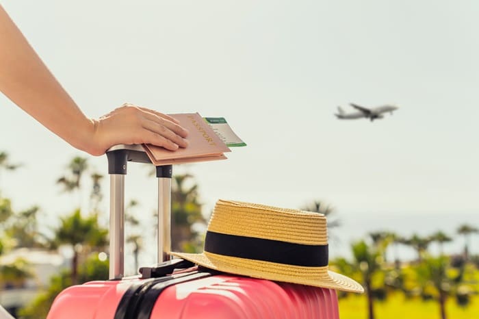 Maringá Turismo dá dicas de destinos para o fim de ano e como economizar nas viagens: foto de uma pessoa, segurando uma mala rosa, com um chapéu apoiado em cima. Ao mesmo tempo, ela segura seu passaporte e passagens na mão direita e olha para um avião